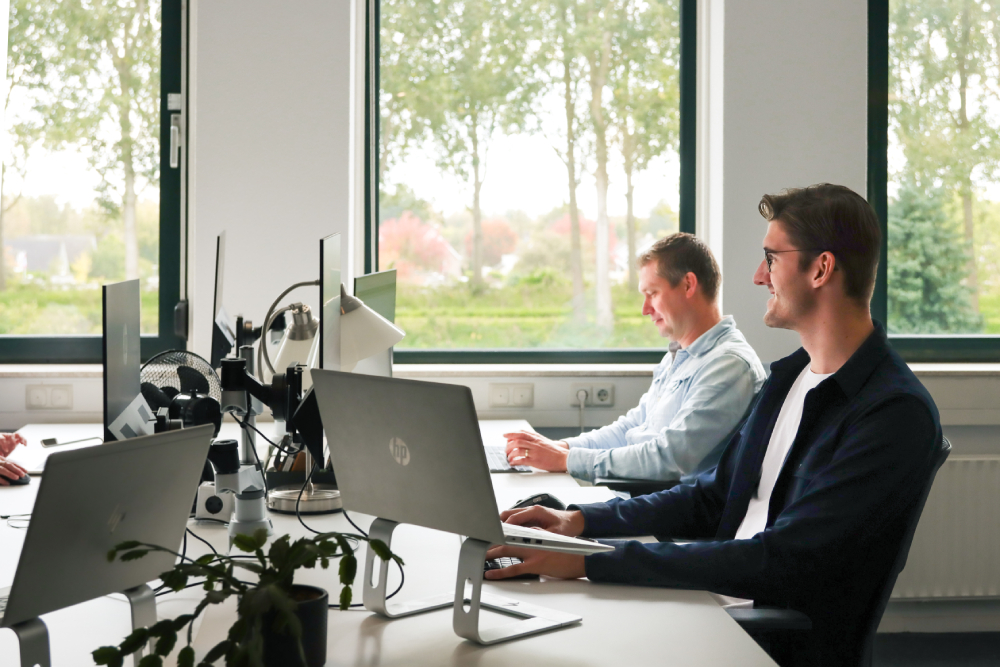 Twee collega’s werken geconcentreerd achter hun computers in een licht kantoor met uitzicht op groen. De sfeer is rustig en gefocust, passend bij efficiënt teamwork en personeelsplanning.