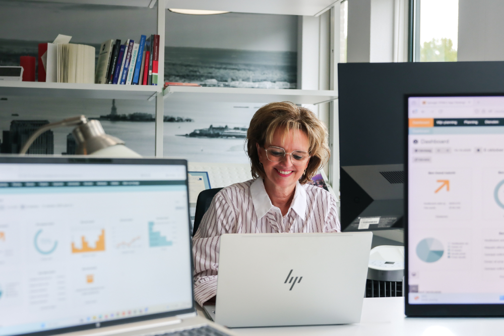 Vrouw werkt glimlachend achter haar laptop, met op de voorgrond schermen vol grafieken en dashboards. De foto benadrukt focus en werkplezier bij datagedreven personeelsplanning.